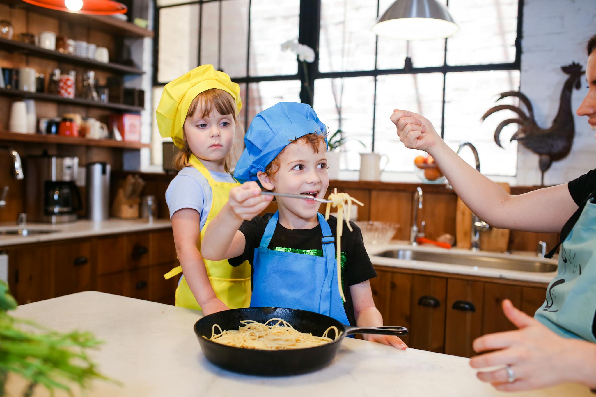 Two children learning to cook spaghetti with guidance in a warm kitchen setting.